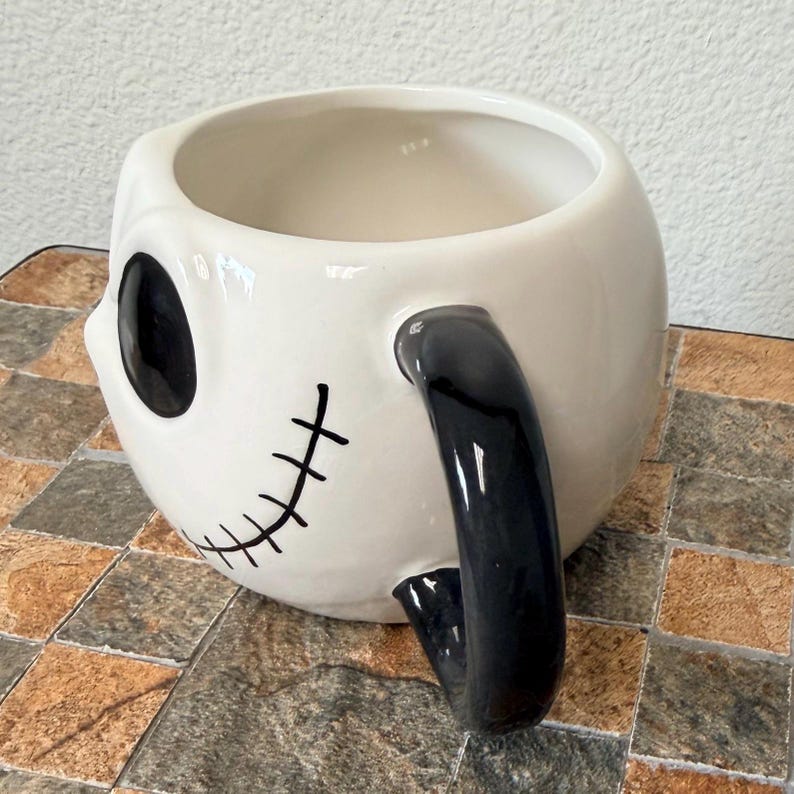 May include: A white ceramic mug with a black handle and skull-like facial features. The mug features a large black eye and a stitched smile. A unique and decorative item.