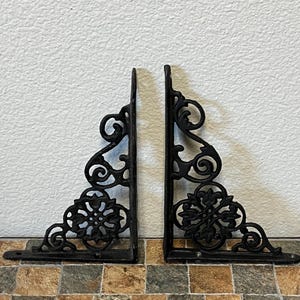 Può includere: Due staffe per mensole in metallo nero ornate. Ogni staffa presenta un design floreale e a volute decorativo. Le staffe sono in ghisa e sono progettate per essere montate a parete.