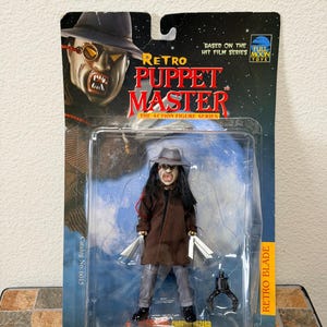 Puede incluir: Figura de acción Retro Puppet Master en un paquete de plástico transparente. La figura lleva un abrigo marrón, pantalones grises y un sombrero. Sostiene dos cuchillas plateadas y una garra negra. El embalaje dice "Retro Puppet Master" y "Retro Blade."