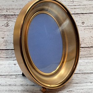 May include: A gold-colored, oval-shaped picture frame with a glass front and a blue background. The frame has a small, round foot for support and a hinged stand on the back.
