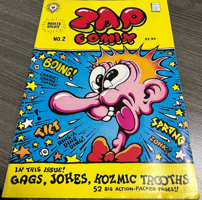 May include: A yellow comic book cover with the title "Zap Comix" in red and black letters. The cover features a cartoon character with a pink face and a large nose. The character is surrounded by stars and the words "Boing!" and "Spring!" in red and black letters. The comic book is labeled "Adults Only!!" and "No. 2".