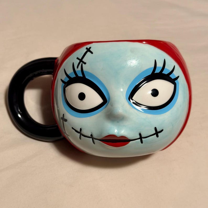 May include: A ceramic mug with a character face design. The mug features a light blue face, red hair, and black stitched details. It has a black handle and a red rim. The character has large white eyes, black eyelashes, and a red mouth.