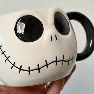 May include: A ceramic mug depicting Jack Skellington's face from The Nightmare Before Christmas. The mug has a white base with black eyes, a stitched mouth, and a black handle. The design is a popular character from the animated film.