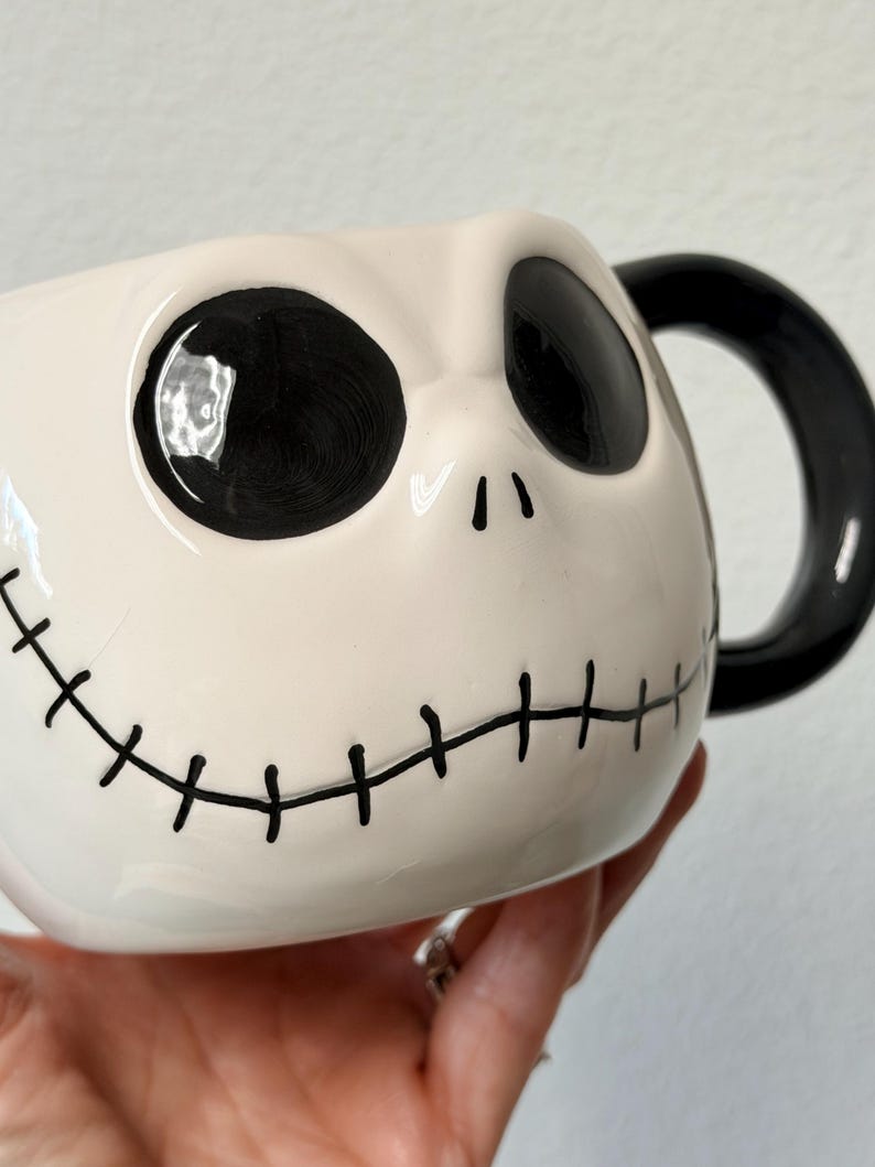 May include: A ceramic mug depicting Jack Skellington's face from The Nightmare Before Christmas. The mug has a white base with black eyes, a stitched mouth, and a black handle. The design is a popular character from the animated film.