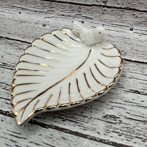May include: A white ceramic leaf-shaped dish with gold trim and a small white bird figurine on top. The dish is perfect for holding jewelry or other small items.