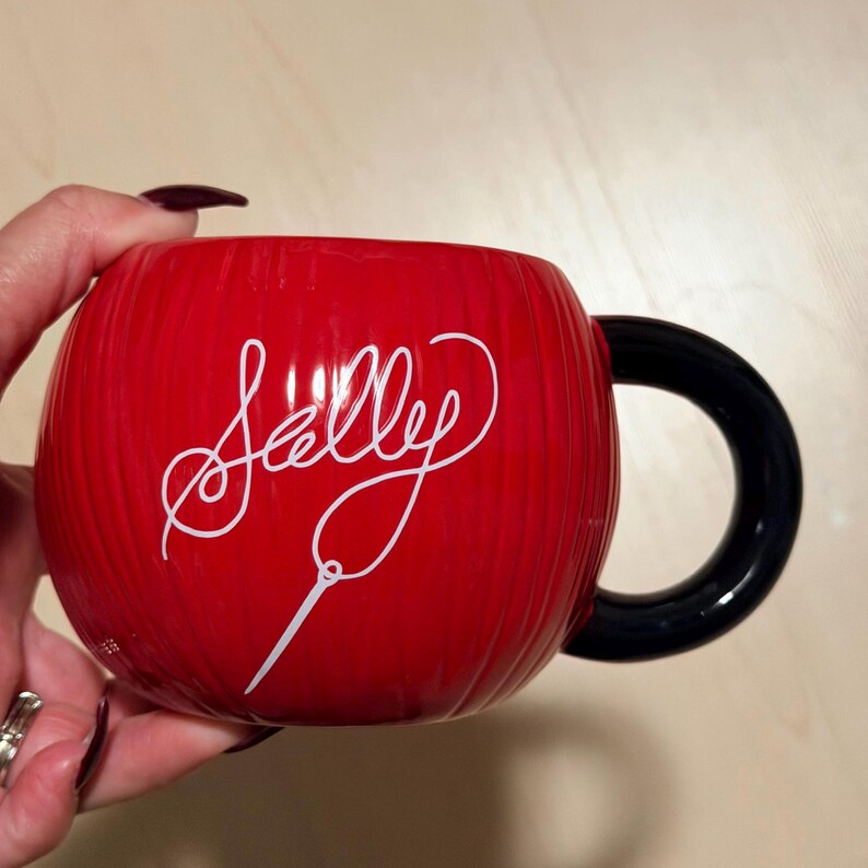 May include: A red ceramic mug with the name "Sally" in white script. The mug has a black handle and a textured exterior. A white needle graphic is below the name. The mug is held by a person.