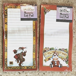 May include: Two Mary Engelbreit magnetic list pads, each with 80 sheets. One features Santa Claus, the other a Christmas character. Both have a red border with gold accents and the text "ONCE A YEAR BUT COMES FOR CHRISTMAS" and "MAKE GOOD CHEER".