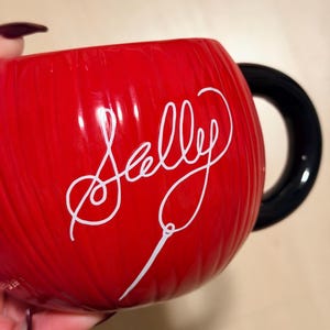 May include: A red ceramic mug with a black handle. The mug features the name "Sally" in white cursive script. The mug has a ribbed texture.