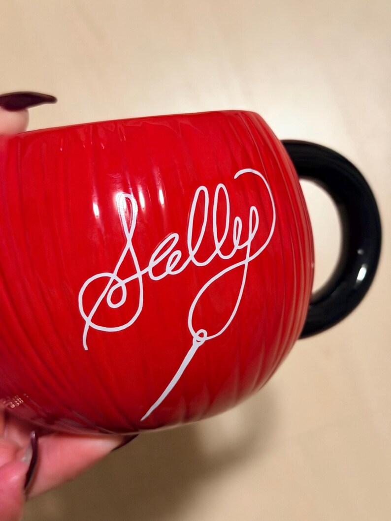 May include: A red ceramic mug with a black handle. The mug features the name "Sally" in white cursive script. The mug has a ribbed texture.