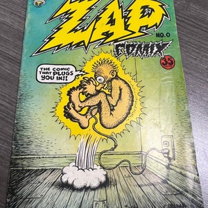May include: A comic book cover with a yellow and black title "ZAP COMIX" and the number "NO. 0". The cover features a cartoon illustration of a nude, humanoid figure with a large eye, plugged into an electrical outlet with a cord. The text "THE COMIC THAT PLUGS YOU IN!!" is in a speech bubble above the figure. The cover is green and yellow.