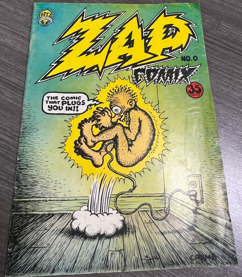 May include: A comic book cover with a yellow and black title "ZAP COMIX" and the number "NO. 0". The cover features a cartoon illustration of a nude, humanoid figure with a large eye, plugged into an electrical outlet with a cord. The text "THE COMIC THAT PLUGS YOU IN!!" is in a speech bubble above the figure. The cover is green and yellow.