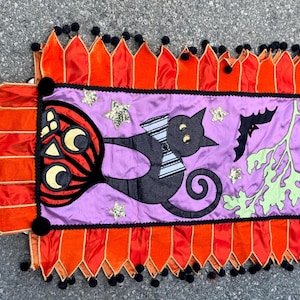 Op de afbeelding: Een rechthoekige stoffen tafelloper met een paars middenpaneel met een zwarte kat met een gestreepte vlinderdas, een pompoen, een vleermuis, een gele halve maan en gouden sterren, afgewerkt met oranje puntige randen en zwarte pompons.