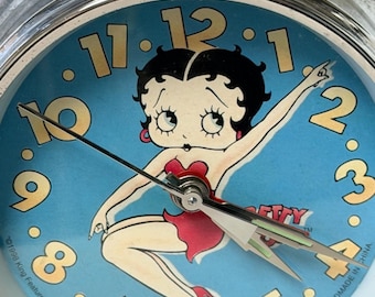 May include: A chrome twin-bell alarm clock featuring a blue face with a central illustration of Betty Boop wearing a red dress. The clock has large yellow numbers, silver hands, and text at the bottom reading 1998 King Features Syndicate, Inc.