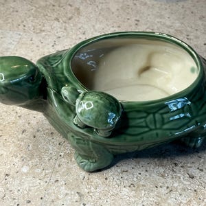 May include: A glossy green ceramic turtle-shaped planter with a cream-colored interior. The turtle features a textured shell pattern and a smaller baby turtle figurine attached to the side of the rim. The piece has four short legs and a head.