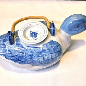 May include: A ceramic duck-shaped teapot with a blue and white design. The teapot features a blue head and a body decorated with blue feather patterns. It has a small, round lid with a floral design and a woven handle.