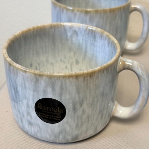 May include: Two ceramic mugs featuring a light blue and white drip glaze pattern with a tan rim. A circular black sticker on the front of the foreground mug displays the text Barcela, Ceramics for Life, Handmade in Portugal, Fait main au Portugal.