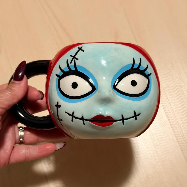 May include: A ceramic mug with a stylized face design, featuring blue skin, black stitched lines, and red hair. The mug has a black handle and is decorated with large eyes, black eyelashes, and a red mouth. A unique piece of drinkware.