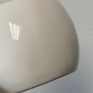 May include: A close-up of a cream-coloured, rounded ceramic object with a visible hairline crack. The surface is smooth and glossy, reflecting light. The object appears to be a part of a larger piece, possibly a mug or a bowl.