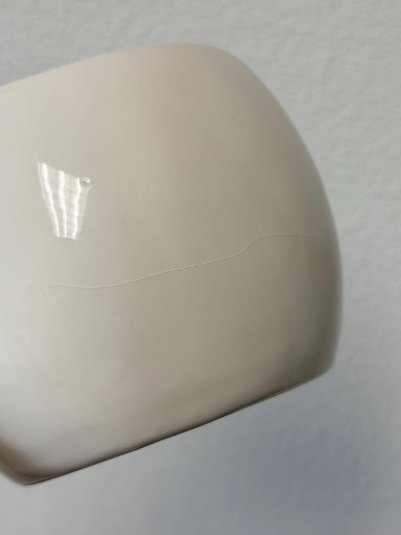 May include: A close-up of a cream-coloured, rounded ceramic object with a visible hairline crack. The surface is smooth and glossy, reflecting light. The object appears to be a part of a larger piece, possibly a mug or a bowl.
