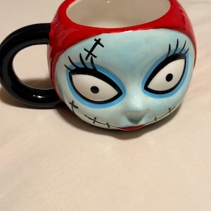 May include: A ceramic mug with a character face design. The mug has blue skin, black stitched details, and large eyes. It features a red rim and a black handle. The design is inspired by a popular animated film.