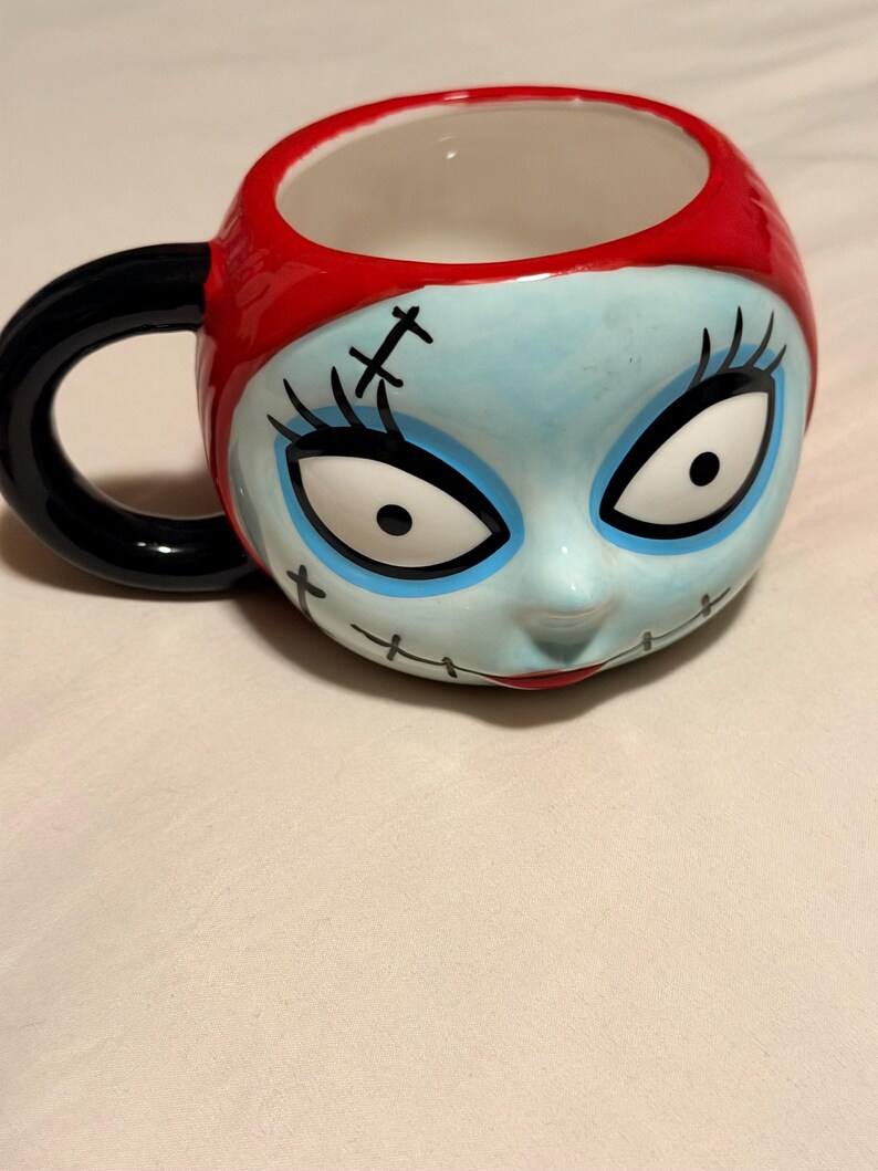 May include: A ceramic mug with a character face design. The mug has blue skin, black stitched details, and large eyes. It features a red rim and a black handle. The design is inspired by a popular animated film.