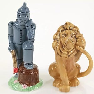 Puede incluir: Dos figuras de cerámica: un hombre de hojalata gris con un hacha y un león marrón. El hombre de hojalata está sentado en un tocón de árbol con una base verde. El león está sentado, con la cola enroscada.