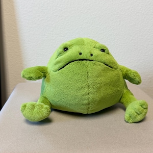 May include: A round, lime green plush frog toy with a soft, fuzzy texture. The stuffed animal features a wide, flat body, two small arms extended to the sides, two legs positioned forward, and a simple embroidered face with two dark eyes.