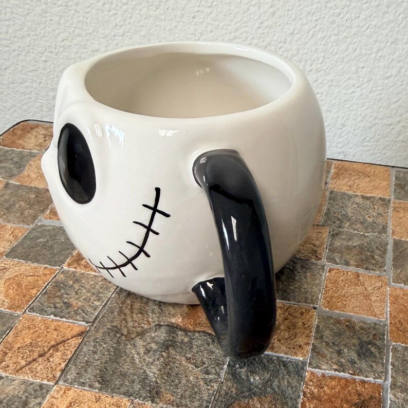 May include: White ceramic mug with a black handle and facial features. The mug has a round shape with a black eye and stitched mouth design. The handle is a curved, solid black. The mug is sitting on a brown and grey tiled surface.