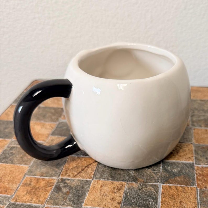May include: A white ceramic mug with a rounded body and a black, glossy handle. The interior of the mug is also white. The mug is sitting on a tiled surface.