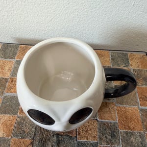 May include: A white ceramic mug shaped like an alien head, with large black oval eyes and a black handle. The inside of the mug is white. The mug is on a brown and tan tiled surface.