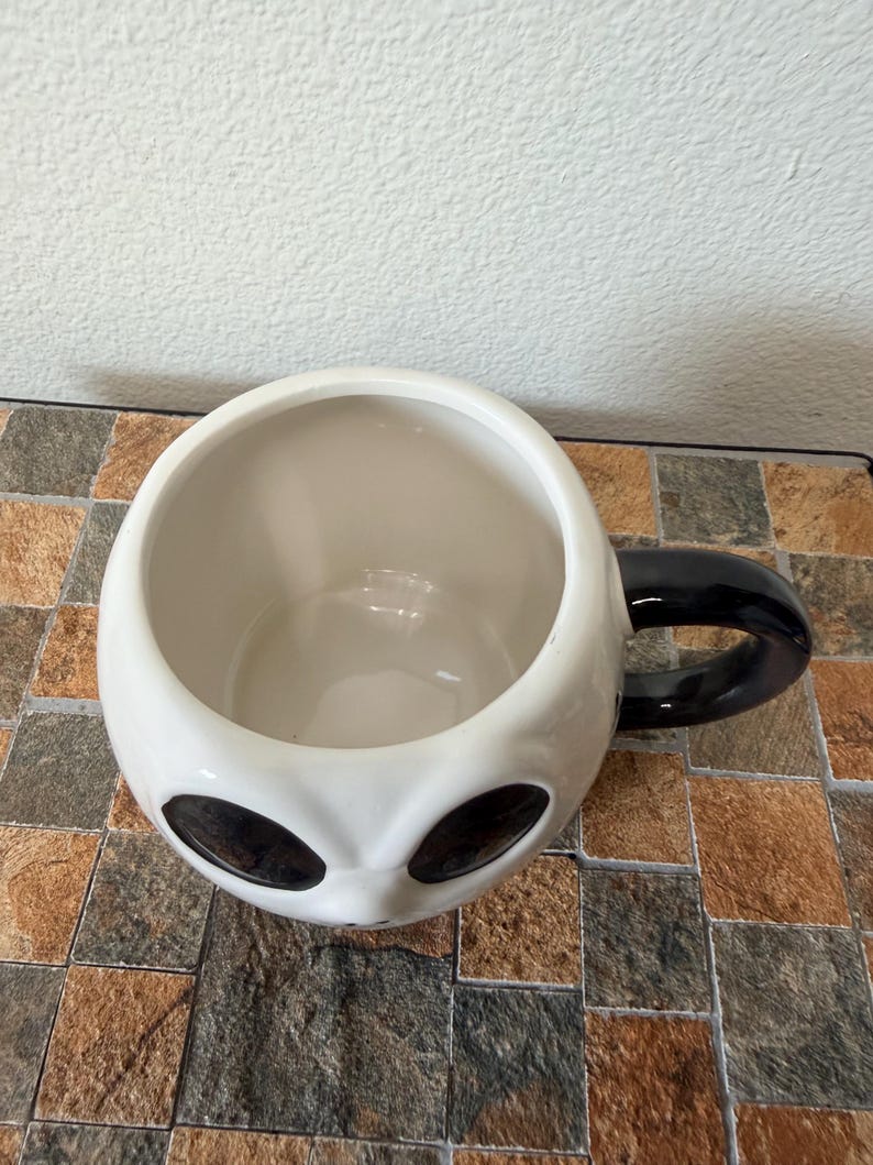 May include: A white ceramic mug shaped like an alien head, with large black oval eyes and a black handle. The inside of the mug is white. The mug is on a brown and tan tiled surface.