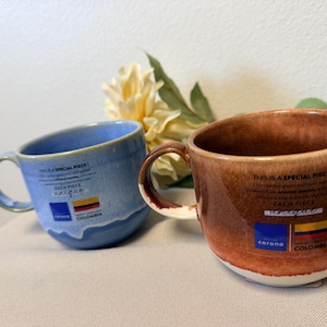 May include: Two ceramic mugs, one blue and one brown, with curved handles. Each mug has text and a flag graphic. The blue mug has a white interior and the brown mug has a white rim. Both mugs are tilted slightly.