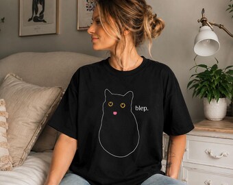 May include: A black short-sleeved t-shirt featuring a white line art illustration of a cat with yellow eyes and a pink tongue sticking out. The text "blep." is printed in white next to the cat graphic on the front of the garment.