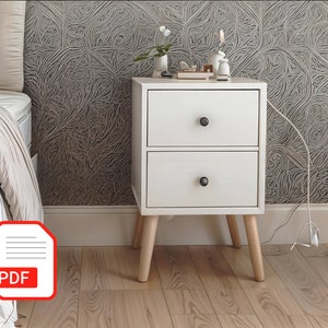 May include: A white two-drawer bedside table with black knobs and light wood legs. The table is next to a bed with a striped duvet and a patterned wall. A lamp and ornaments sit on top.