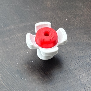 May include: A red and white bottle stopper. The stopper has a cylindrical red top with a hole in the centre, surrounded by white, petal-shaped supports. The design is a cross shape.