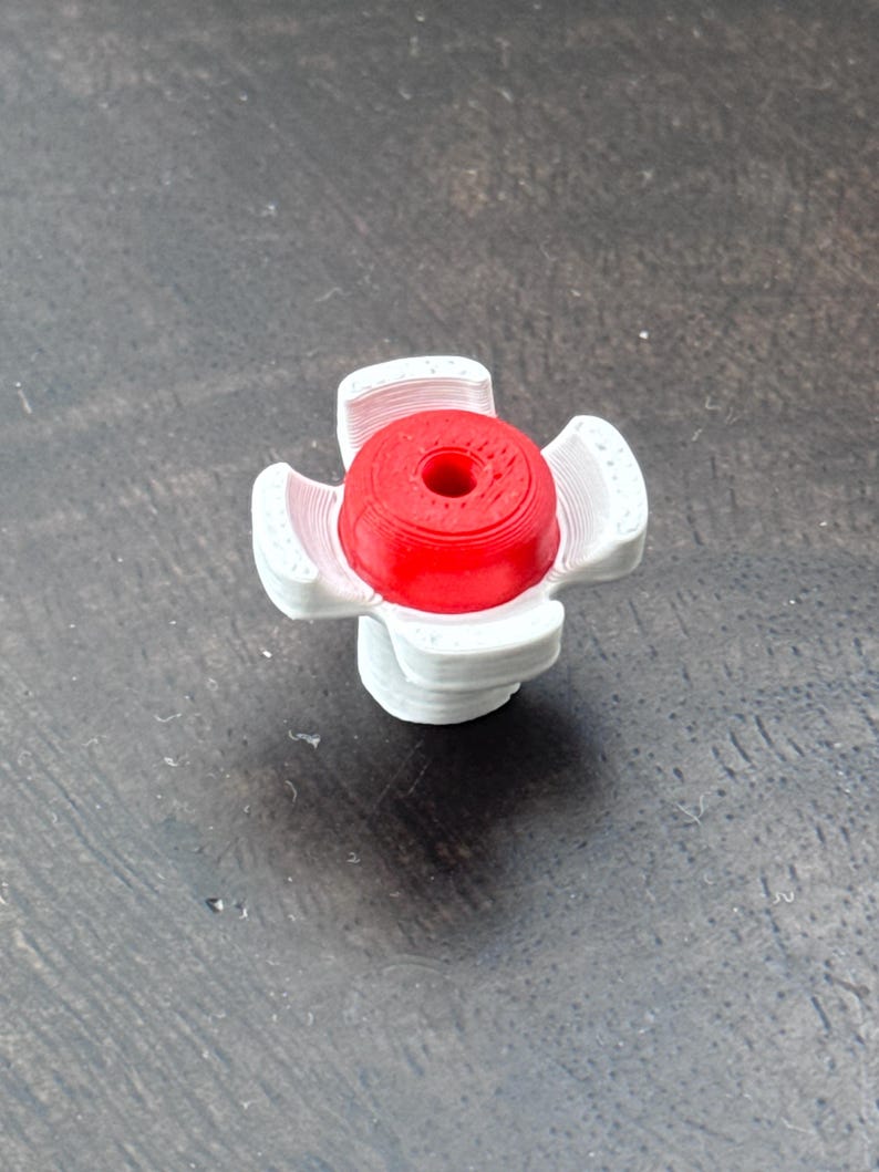 May include: A red and white bottle stopper. The stopper has a cylindrical red top with a hole in the centre, surrounded by white, petal-shaped supports. The design is a cross shape.