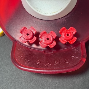 May include: Close-up of a red plastic hummingbird feeder base with three red flower-shaped feeding ports. The base has a clear, white container and bird footstep details.