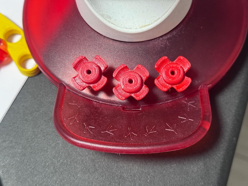 May include: Close-up of a red plastic hummingbird feeder base with three red flower-shaped feeding ports. The base has a clear, white container and bird footstep details.