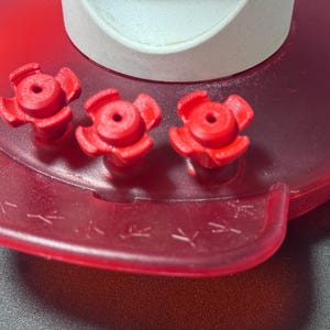 May include: Close-up of a red plastic chicken waterer. The waterer has three red flower-shaped valves and a red base with a small trough. The top is white. The trough has small chicken foot prints.