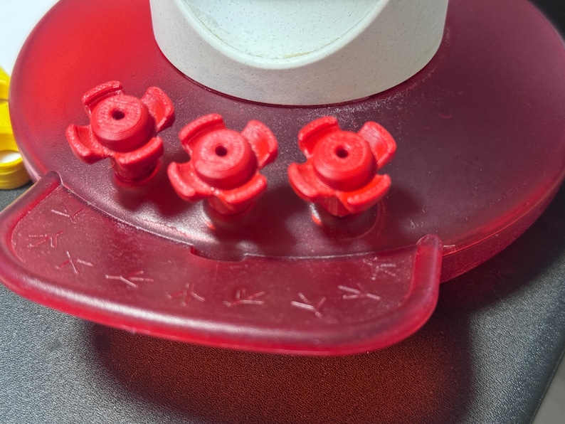 May include: Close-up of a red plastic chicken waterer. The waterer has three red flower-shaped valves and a red base with a small trough. The top is white. The trough has small chicken foot prints.