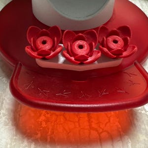 May include: A red hummingbird feeder with three red flower-shaped feeding ports. The feeder has a circular base and a clear, textured bottom. The design includes small bird silhouettes.