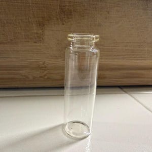 Könnte beinhalten: Ein durchsichtiges Glasfläschchen, zylindrisch geformt mit rundem Boden und schmaler Öffnung. Das Fläschchen ist leer. Der Hintergrund zeigt eine Holzoberfläche und weiße Fliesen. Ideal zur Aufbewahrung von Flüssigkeiten.