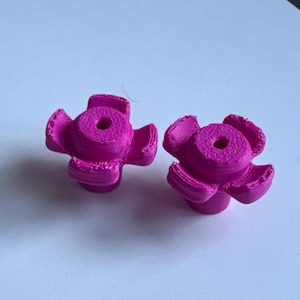 May include: Two bright magenta, flower-shaped cabinet knobs. Each knob has a central circular body with a hole and five curved petals. The knobs are made of a 3D-printed material.