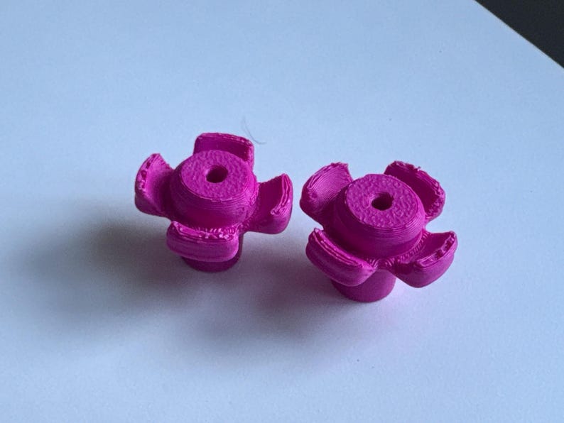 May include: Two bright magenta, flower-shaped cabinet knobs. Each knob has a central circular body with a hole and five curved petals. The knobs are made of a 3D-printed material.
