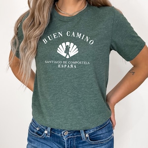 May include: A heathered dark green short-sleeve t-shirt featuring a white graphic of a scallop shell with a hiker silhouette inside. The text reads BUEN CAMINO, SANTIAGO DE COMPOSTELA, ESPAÑA in a white serif font centered on the chest area.
