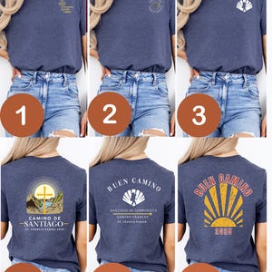 May include: A collection of navy blue t-shirts with various designs. Some shirts feature text and graphics related to the Camino de Santiago pilgrimage, including a cross, scallop shell, and the phrase "Buen Camino". The shirts are displayed on a person.