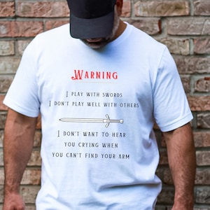 Puede incluir: Camiseta blanca con una ilustración en blanco y negro de una espada y el texto "WARNING I PLAY WITH SWORDS I DON'T PLAY WELL WITH OTHERS I DON'T WANT TO HEAR YOU CRYING WHEN YOU CAN'T FIND YOUR ARM"