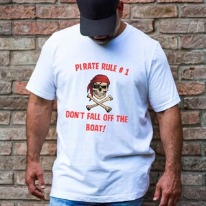 May include: White t-shirt featuring a pirate skull with a red bandana and crossed bones. The text "PIRATE RULE #1" and "DON'T FALL OFF THE BOAT!" are printed in red. The shirt is worn by a person.