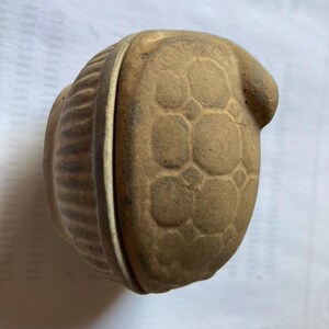 Op de afbeelding: Een kleine, ronde, beige keramische pot met deksel en een textuurrijk oppervlak. Het deksel heeft een verhoogd, geometrisch honingraatpatroon, terwijl de onderkant van de pot is voorzien van verticale, geribbelde details.