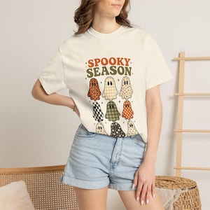 May include: A cream-coloured short-sleeve t-shirt featuring the text SPOOKY SEASON in orange and green letters. Below the text are nine ghost illustrations in various patterns including leopard print, plaid, polka dots, and floral designs.